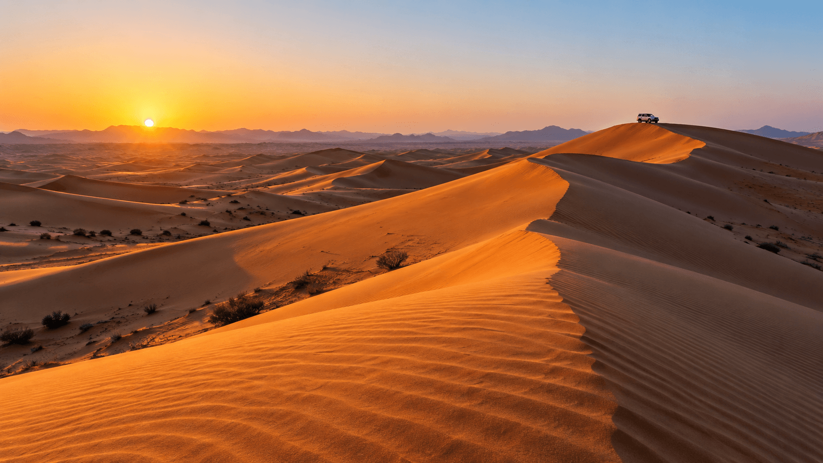 Desert dunes glowing orange at sunrise with a 4x4 parked on a ridge